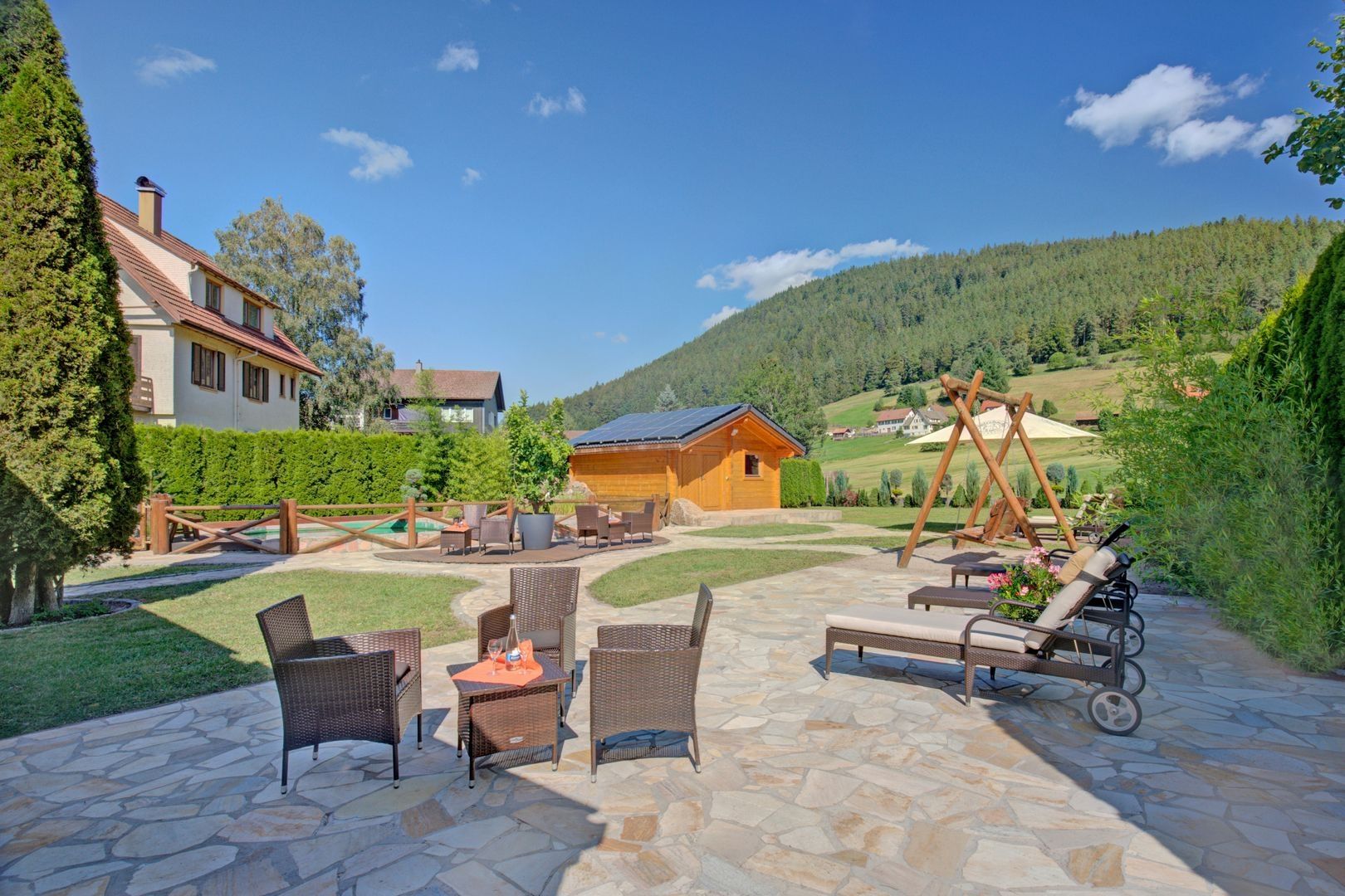 Sunny outdoor terrace with wicker chairs and sun loungers on stone tiles. Lush green hills and a wooden cabin.