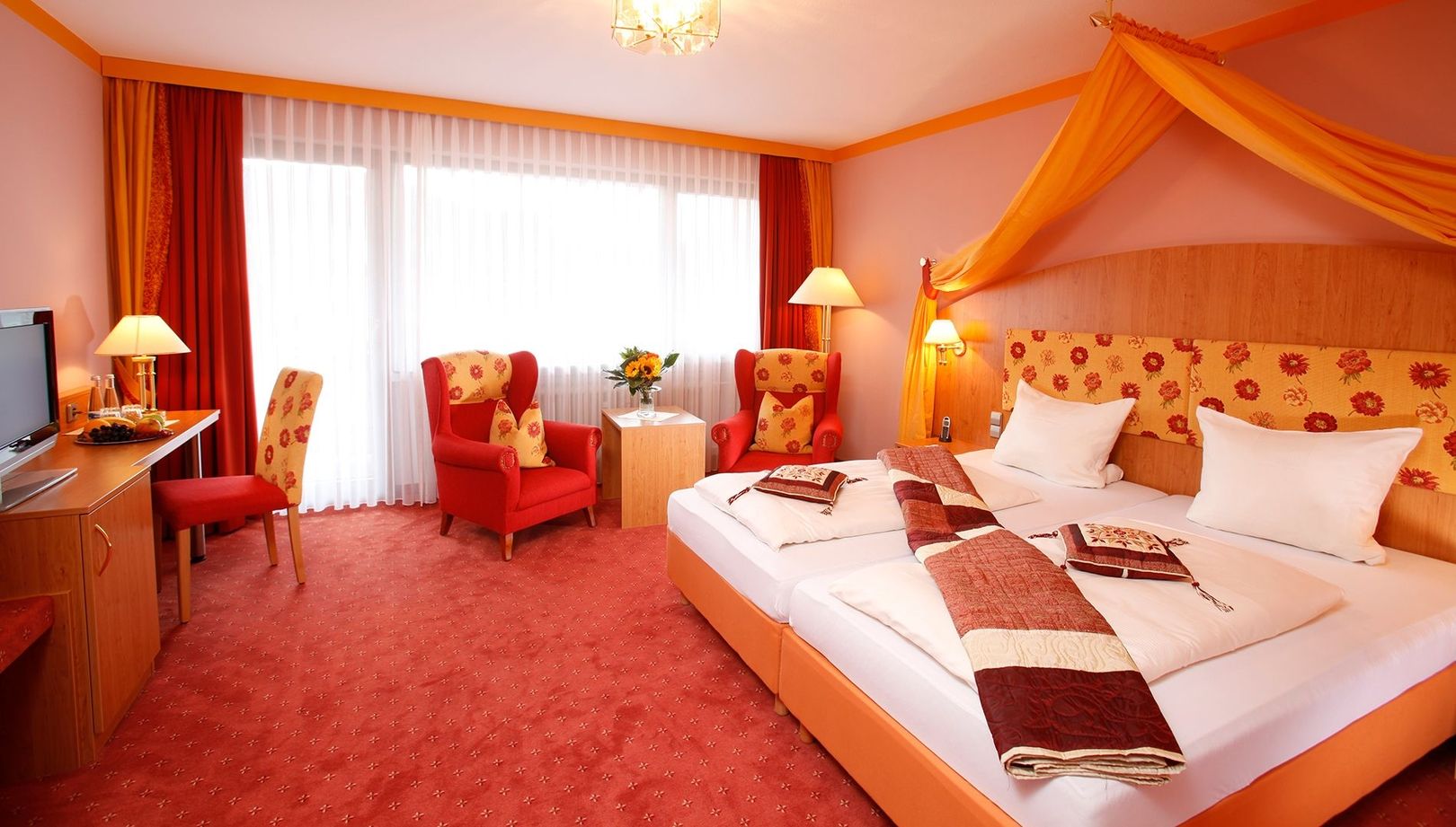 Une chambre d'hôtel confortable est dotée d'un lit double aux draps blancs à motifs, encadré de rideaux jaune chaud. Deux fauteuils rouges sont placés près de la fenêtre.