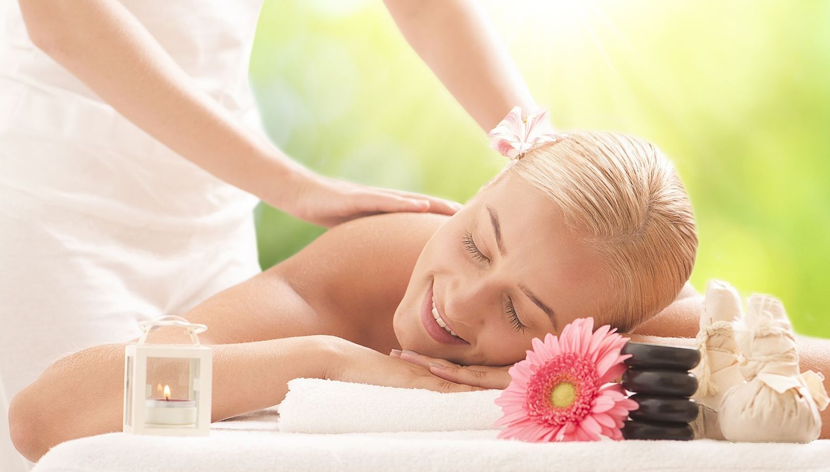 A woman enjoys a relaxing massage, lying on her stomach and smiling serenely. Near her are black stones, a pink flower, a burning candle, and herbal sachets.