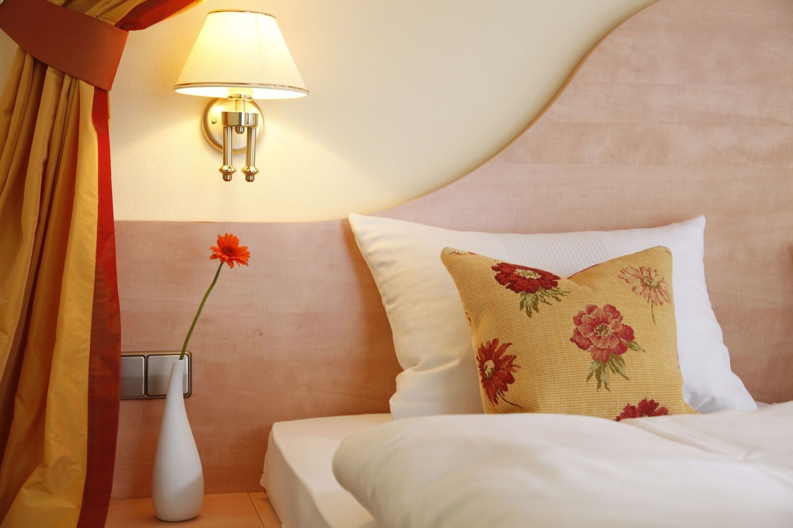 Un coin douillet dans la chambre, avec un lit orné d'oreillers blancs et d'un coussin à motifs floraux. Une applique murale diffuse une lumière douce, et un vase contenant une fleur orange apporte une touche d'élégance.
