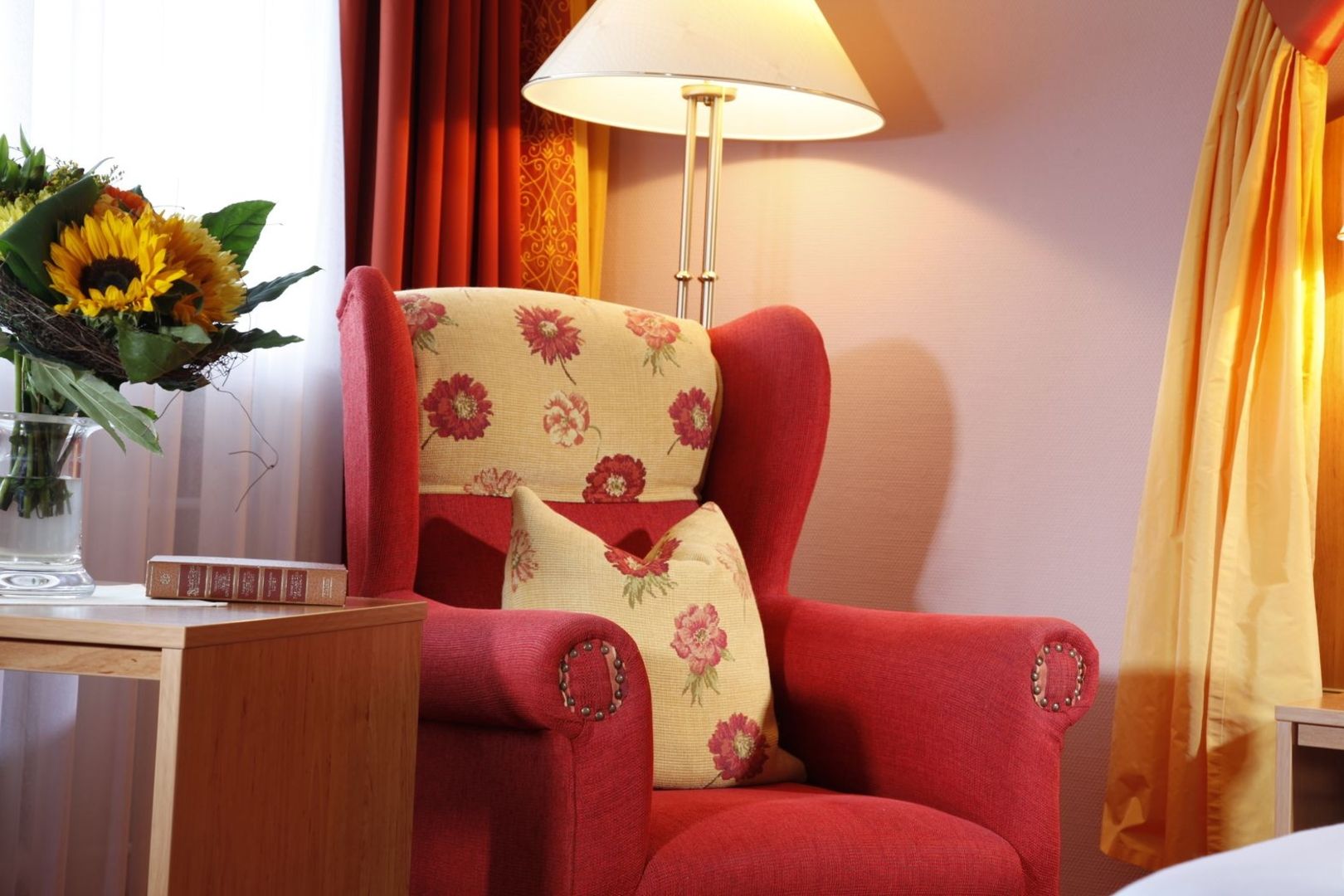 Cozy room with a red armchair with a floral pattern, a bouquet of sunflowers on a side table, warm yellow and red curtains, and a burning lamp.